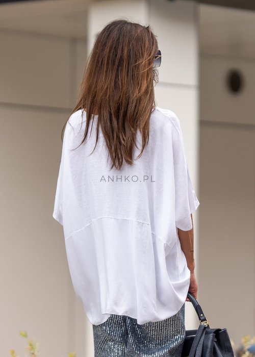 Biała bluzka oversize z lnem o szerokim kroju i krótkim rękawie, z dłuższym tyłem oraz subtelnym łączeniem materiałów. Model pokazany w stylizacji z połyskującymi spodniami i czarną torebką.