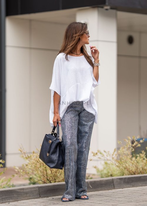 Biała bluzka oversize z lnem o szerokim kroju i krótkim rękawie, z dłuższym tyłem oraz subtelnym łączeniem materiałów. Model pokazany w stylizacji z połyskującymi spodniami i czarną torebką.