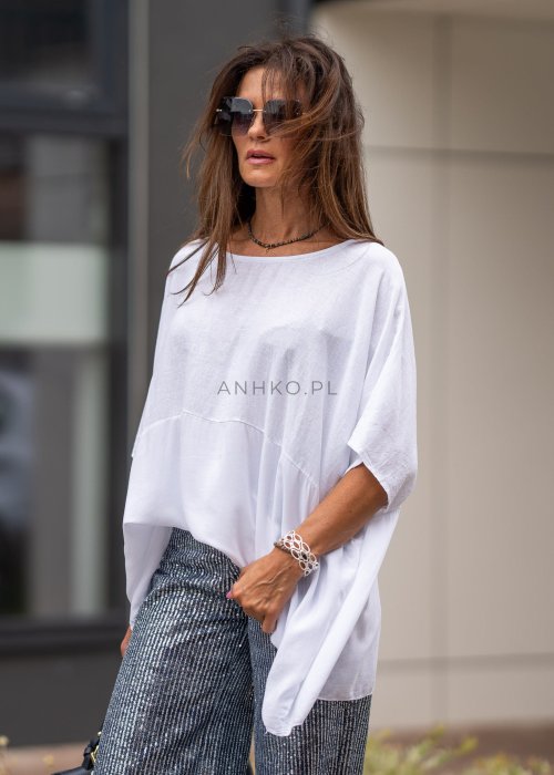 Biała bluzka oversize z lnem o szerokim kroju i krótkim rękawie, z dłuższym tyłem oraz subtelnym łączeniem materiałów. Model pokazany w stylizacji z połyskującymi spodniami i czarną torebką.