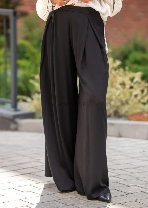 Eleganckie spodnie damskie w kolorze czarnym, z wysokim stanem, szerokimi nogawkami typu palazzo, zakładkami przy talii i asymetrycznym, kopertowym wykończeniem pasa.