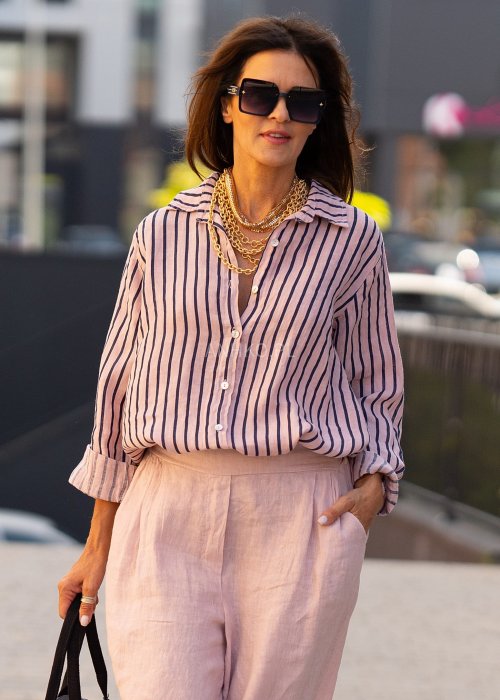 Lniana koszula damska w różowym kolorze z granatowymi paskami, luźny fason z kołnierzykiem i guzikami, stylizacja z jasnymi spodniami.