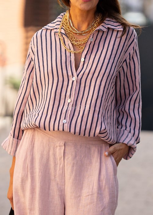 Lniana koszula damska w różowym kolorze z granatowymi paskami, luźny fason z kołnierzykiem i guzikami, stylizacja z jasnymi spodniami.