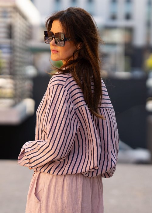 Lniana koszula damska w różowym kolorze z granatowymi paskami, luźny fason z kołnierzykiem i guzikami, stylizacja z jasnymi spodniami.