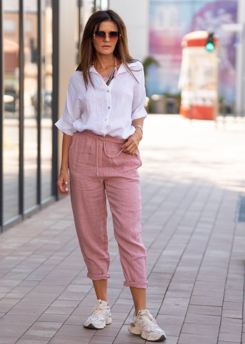 Jasnoróżowe lniane spodnie damskie z gumą w pasie i kieszeniami, model o swobodnym kroju stylizowany z białą koszulą i sneakersami.