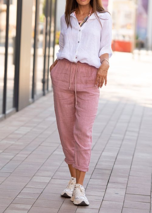 Jasnoróżowe lniane spodnie damskie z gumą w pasie i kieszeniami, model o swobodnym kroju stylizowany z białą koszulą i sneakersami.