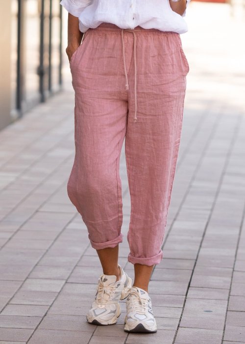 Jasnoróżowe lniane spodnie damskie z gumą w pasie i kieszeniami, model o swobodnym kroju stylizowany z białą koszulą i sneakersami.