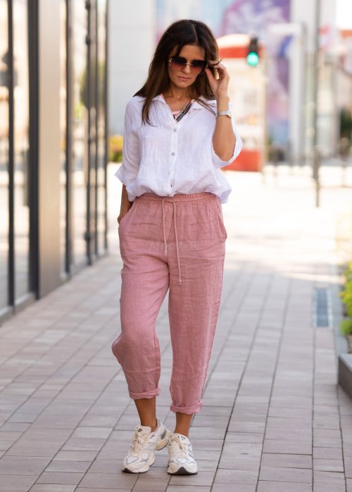 Jasnoróżowe lniane spodnie damskie z gumą w pasie i kieszeniami, model o swobodnym kroju stylizowany z białą koszulą i sneakersami.