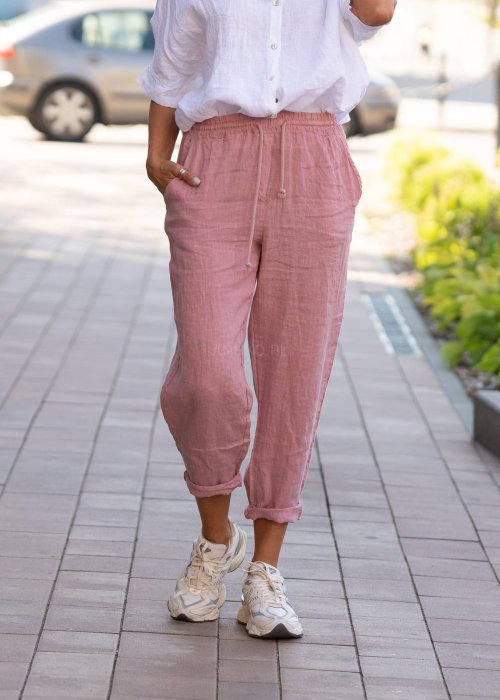 Jasnoróżowe lniane spodnie damskie z gumą w pasie i kieszeniami, model o swobodnym kroju stylizowany z białą koszulą i sneakersami.