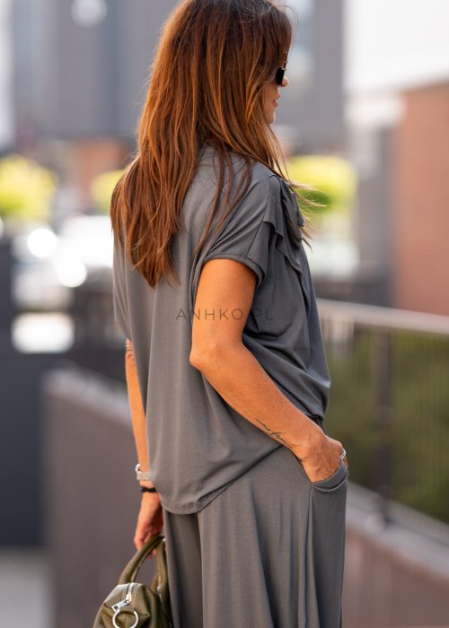 Grafitowa bluzka damska o luźnym kroju z dekoltem w łódkę i materiałowym kwiatem na ramieniu. Stylizacja z szerokimi spodniami w eleganckim, miejskim stylu.