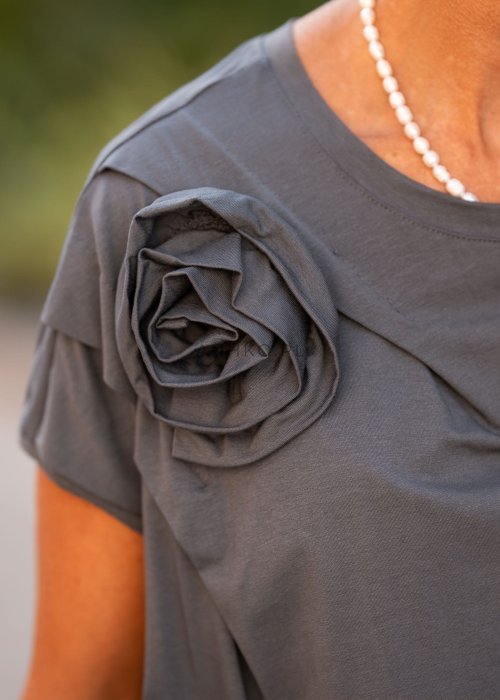 Grafitowa bluzka damska o luźnym kroju z dekoltem w łódkę i materiałowym kwiatem na ramieniu. Stylizacja z szerokimi spodniami w eleganckim, miejskim stylu.