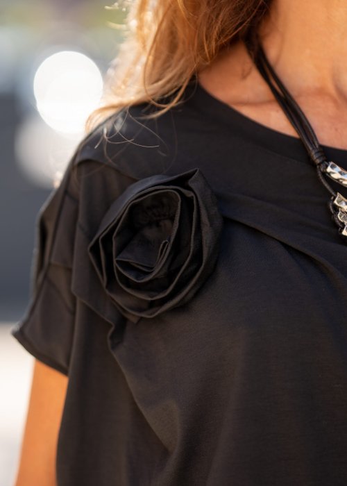 Czarna bluzka damska o luźnym kroju z dekoltem w łódkę i dużym materiałowym kwiatem na ramieniu. Model zestawiony z szerokimi czarnymi spodniami w eleganckiej, miejskiej stylizacji.