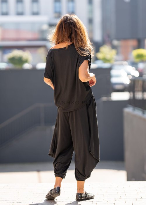 Czarna bluzka damska o luźnym kroju z dekoltem w łódkę i dużym materiałowym kwiatem na ramieniu. Model zestawiony z szerokimi czarnymi spodniami w eleganckiej, miejskiej stylizacji.