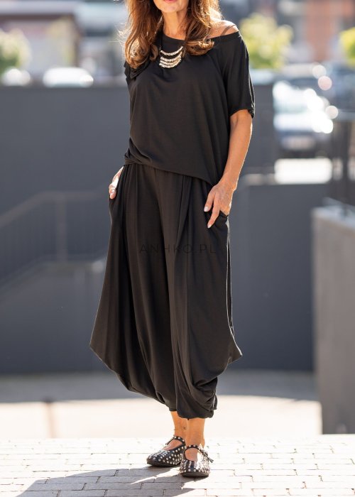 Czarna bluzka damska o luźnym kroju z dekoltem w łódkę i dużym materiałowym kwiatem na ramieniu. Model zestawiony z szerokimi czarnymi spodniami w eleganckiej, miejskiej stylizacji.