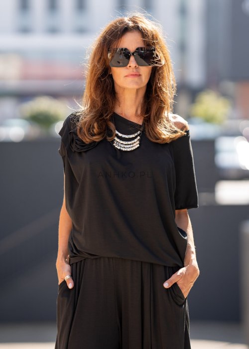 Czarna bluzka damska o luźnym kroju z dekoltem w łódkę i dużym materiałowym kwiatem na ramieniu. Model zestawiony z szerokimi czarnymi spodniami w eleganckiej, miejskiej stylizacji.