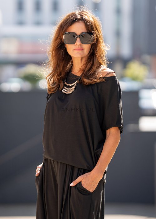 Czarna bluzka damska o luźnym kroju z dekoltem w łódkę i dużym materiałowym kwiatem na ramieniu. Model zestawiony z szerokimi czarnymi spodniami w eleganckiej, miejskiej stylizacji.