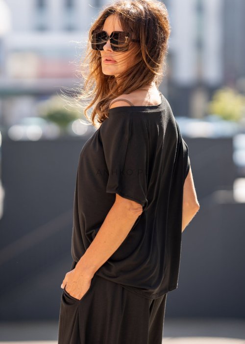 Czarna bluzka damska o luźnym kroju z dekoltem w łódkę i dużym materiałowym kwiatem na ramieniu. Model zestawiony z szerokimi czarnymi spodniami w eleganckiej, miejskiej stylizacji.