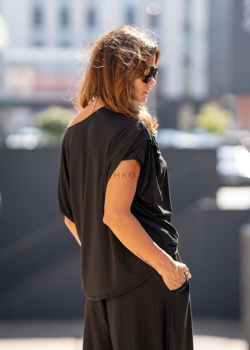 Czarna bluzka damska o luźnym kroju z dekoltem w łódkę i dużym materiałowym kwiatem na ramieniu. Model zestawiony z szerokimi czarnymi spodniami w eleganckiej, miejskiej stylizacji.