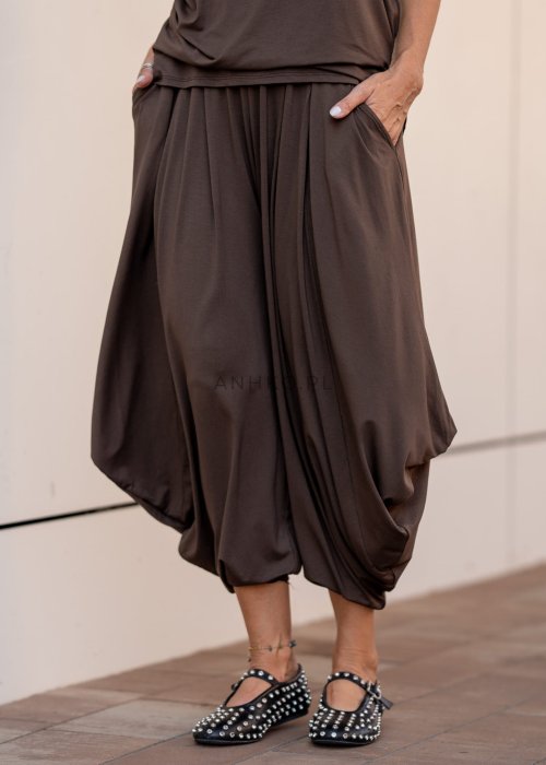 Brązowe spodnie damskie typu harem z szerokimi, drapowanymi nogawkami i elastycznym pasem. Stylizacja z luźnym topem i skórzanymi butami z ćwiekami.