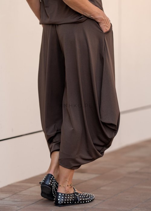 Brązowe spodnie damskie typu harem z szerokimi, drapowanymi nogawkami i elastycznym pasem. Stylizacja z luźnym topem i skórzanymi butami z ćwiekami.