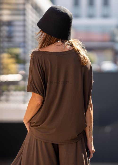 Brązowa bluzka damska o luźnym kroju z dekoltem w łódkę i materiałowym kwiatem na ramieniu. Stylizacja z szerokimi spodniami w eleganckim, miejskim stylu.