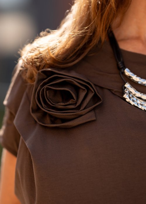 Brązowa bluzka damska o luźnym kroju z dekoltem w łódkę i materiałowym kwiatem na ramieniu. Stylizacja z szerokimi spodniami w eleganckim, miejskim stylu.