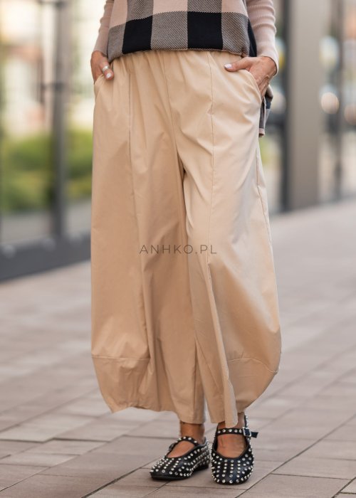 Beżowe spodnie damskie o szerokich nogawkach z efektem bombki i gumką w pasie, zestawione z czarnymi butami z ćwiekami w miejskiej stylizacji.