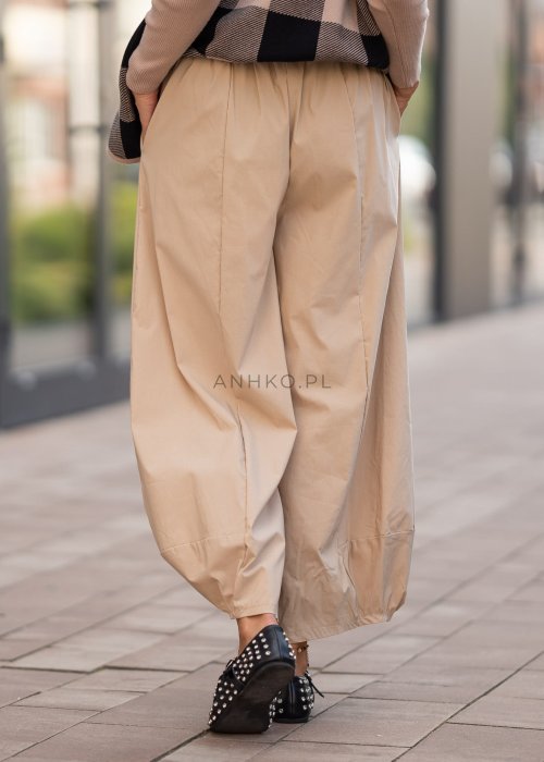 Beżowe spodnie damskie o szerokich nogawkach z efektem bombki i gumką w pasie, zestawione z czarnymi butami z ćwiekami w miejskiej stylizacji.