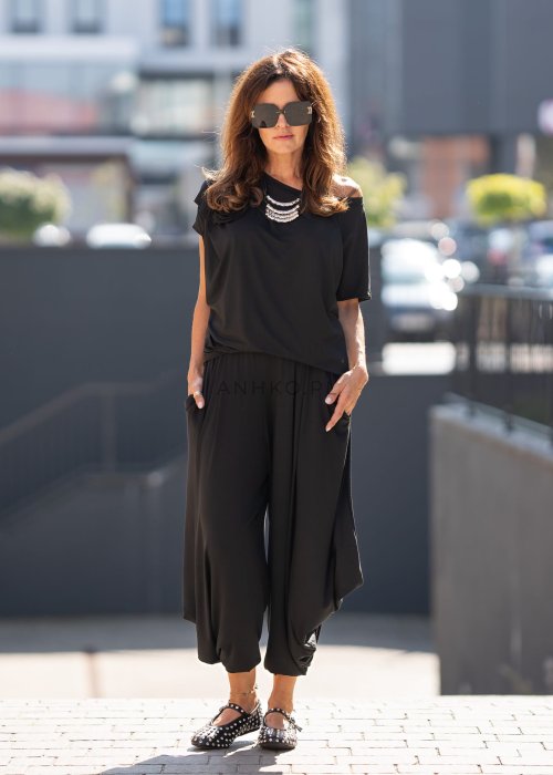 Czarna bluzka damska o luźnym kroju z dekoltem w łódkę i dużym materiałowym kwiatem na ramieniu. Model zestawiony z szerokimi czarnymi spodniami w eleganckiej, miejskiej stylizacji.
