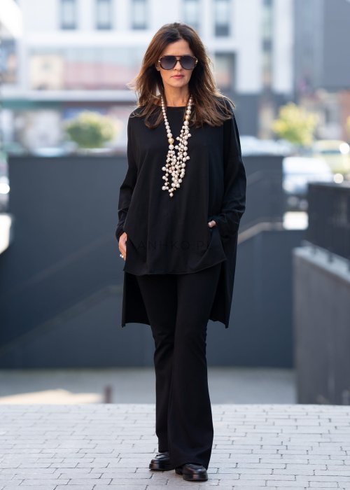 Czarna bluzka oversize z asymetrycznym dołem i dłuższym tyłem, zestawiona z czarnymi spodniami w eleganckiej, miejskiej stylizacji.