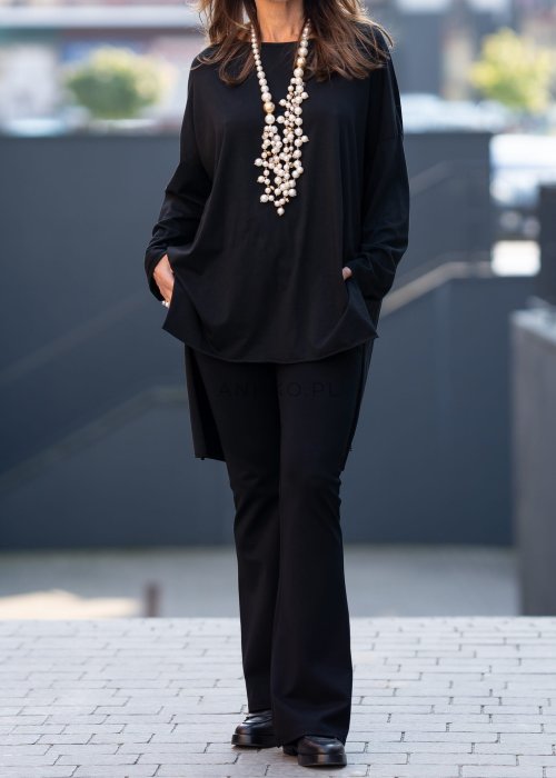 Czarna bluzka oversize z asymetrycznym dołem i dłuższym tyłem, zestawiona z czarnymi spodniami w eleganckiej, miejskiej stylizacji.