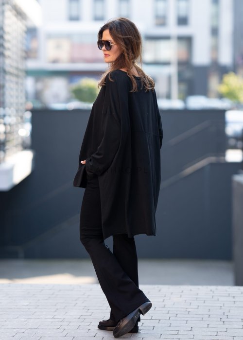 Czarna bluzka oversize z asymetrycznym dołem i dłuższym tyłem, zestawiona z czarnymi spodniami w eleganckiej, miejskiej stylizacji.