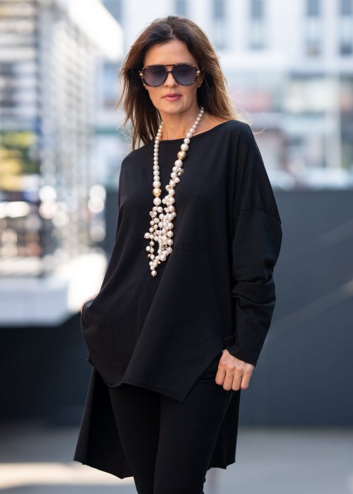 Czarna bluzka oversize z asymetrycznym dołem i dłuższym tyłem, zestawiona z czarnymi spodniami w eleganckiej, miejskiej stylizacji.