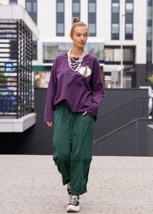 Damskie spodnie dresowe w kolorze butelkowej zieleni o luźnym kroju z ozdobnymi przeszyciami i kieszeniami. Stylizacja z podwiniętymi nogawkami i sneakersami.