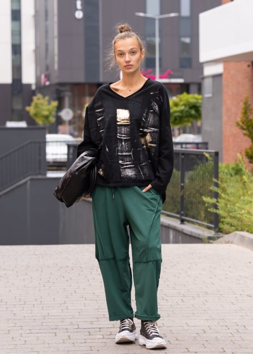 Damskie spodnie dresowe w kolorze butelkowej zieleni o luźnym kroju z ozdobnymi przeszyciami i kieszeniami. Stylizacja z podwiniętymi nogawkami i sneakersami.