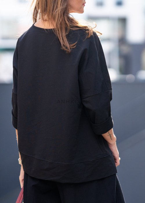 Czarna bluza damska oversize z asymetrycznym dołem i przeszyciami. Model prezentowany w minimalistycznej stylizacji z szerokimi spodniami i biżuterią.