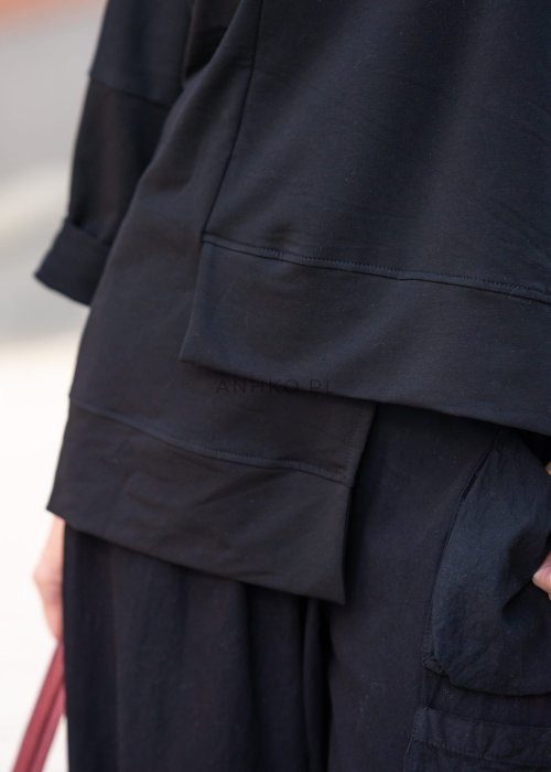 Czarna bluza damska oversize z asymetrycznym dołem i przeszyciami. Model prezentowany w minimalistycznej stylizacji z szerokimi spodniami i biżuterią.