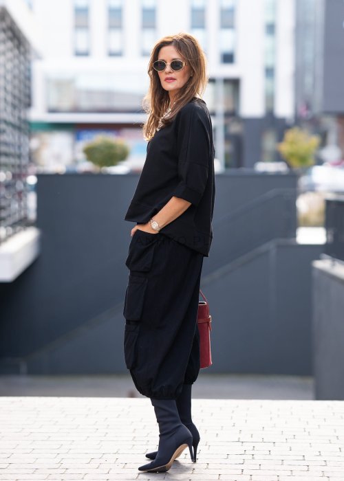 Czarna bluza damska oversize z asymetrycznym dołem i przeszyciami. Model prezentowany w minimalistycznej stylizacji z szerokimi spodniami i biżuterią.