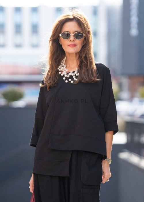 Czarna bluza damska oversize z asymetrycznym dołem i przeszyciami. Model prezentowany w minimalistycznej stylizacji z szerokimi spodniami i biżuterią.