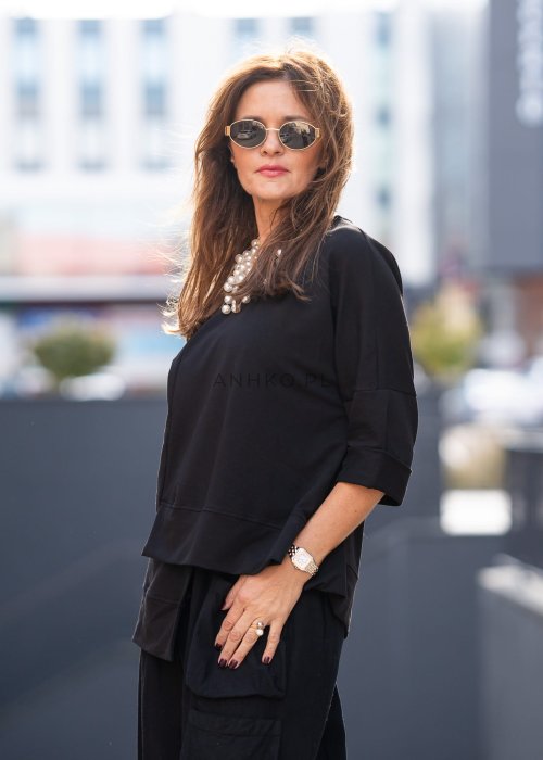 Czarna bluza damska oversize z asymetrycznym dołem i przeszyciami. Model prezentowany w minimalistycznej stylizacji z szerokimi spodniami i biżuterią.