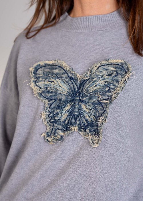 Dzianinowy sweter damski w jasnoszarym kolorze, o luźnym fasonie z długimi rękawami i dekoracyjną aplikacją motyla z denimu.