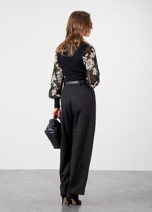 Czarne spodnie damskie z wysokim stanem, zakładkami i paskiem z metalową klamrą. Elegancki model z szeroką nogawką pokazany w stylizacji z dopasowaną górą i botkami na obcasie.