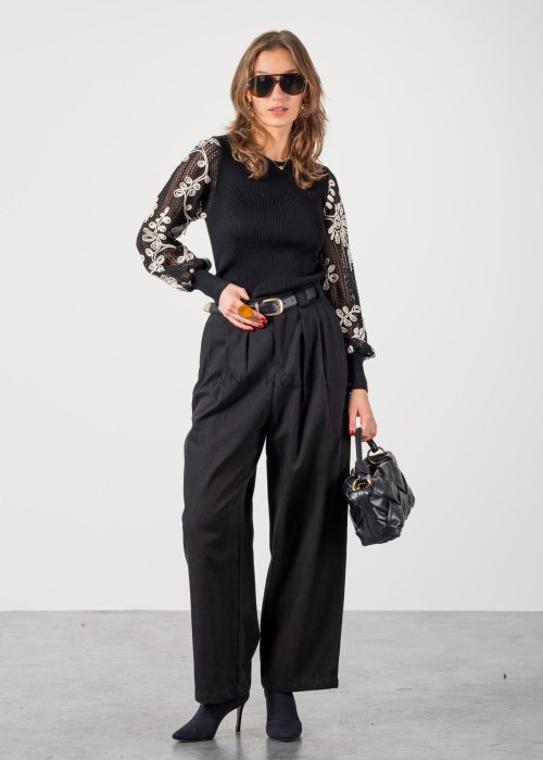 Czarne spodnie damskie z wysokim stanem, zakładkami i paskiem z metalową klamrą. Elegancki model z szeroką nogawką pokazany w stylizacji z dopasowaną górą i botkami na obcasie.