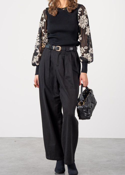 Czarne spodnie damskie z wysokim stanem, zakładkami i paskiem z metalową klamrą. Elegancki model z szeroką nogawką pokazany w stylizacji z dopasowaną górą i botkami na obcasie.