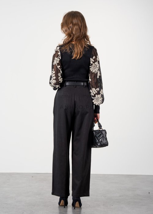 Czarne spodnie damskie z wysokim stanem, zakładkami i paskiem z metalową klamrą. Elegancki model z szeroką nogawką pokazany w stylizacji z dopasowaną górą i botkami na obcasie.