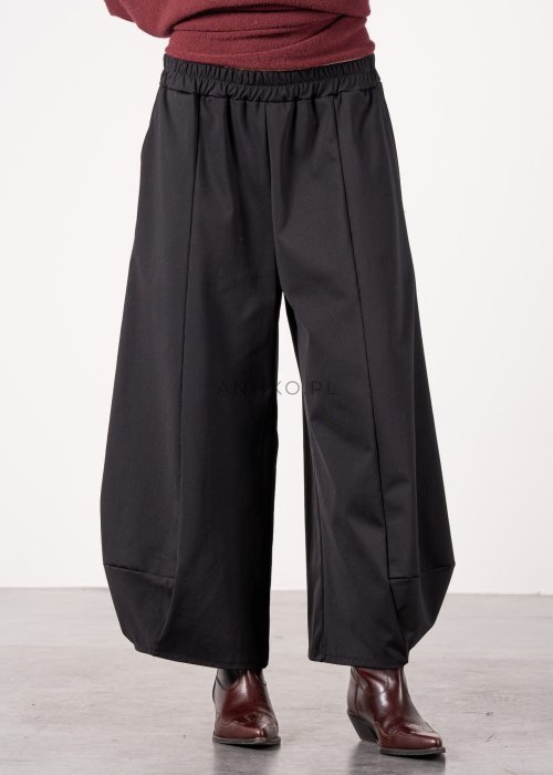 Czarne spodnie damskie o szerokich nogawkach z efektem bombki i elastyczną talią, zestawione z botkami w minimalistycznej stylizacji.