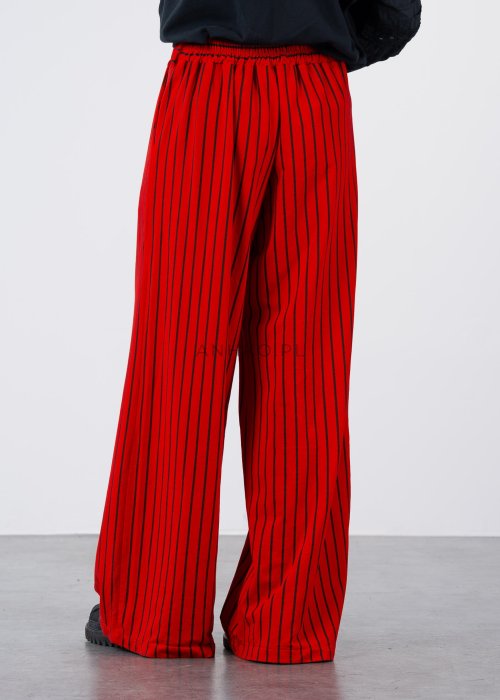 Czerwone spodnie damskie w czarne pionowe paski o szerokim kroju, zestawione z czarną bluzą i botkami na obcasie w nowoczesnej stylizacji.