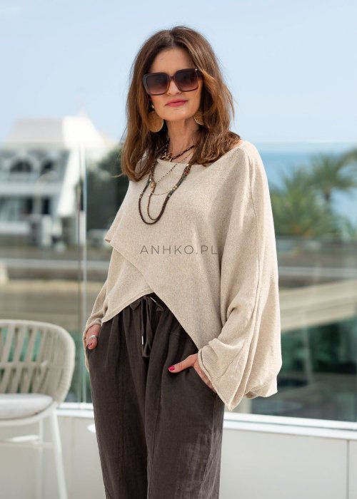 Kremowy sweter oversize z asymetrycznym dołem i szerokimi rękawami, zestawiony z brązowymi spodniami o luźnym kroju. Stylizacja w klimacie casual premium z botkami na obcasie.