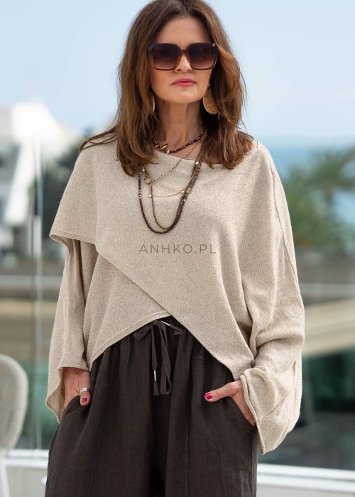 Kremowy sweter oversize z asymetrycznym dołem i szerokimi rękawami, zestawiony z brązowymi spodniami o luźnym kroju. Stylizacja w klimacie casual premium z botkami na obcasie.