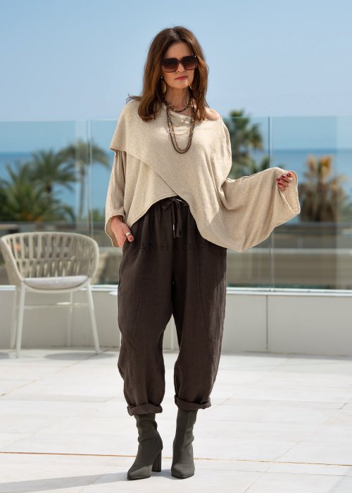 Kremowy sweter oversize z asymetrycznym dołem i szerokimi rękawami, zestawiony z brązowymi spodniami o luźnym kroju. Stylizacja w klimacie casual premium z botkami na obcasie.