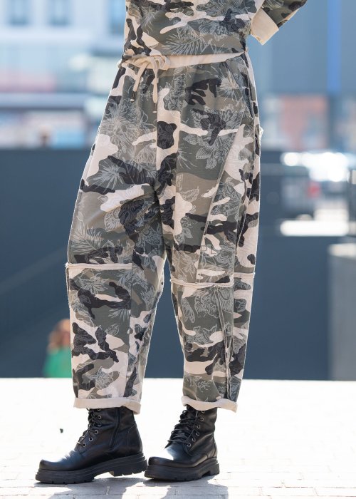 Damskie spodnie dresowe w print moro z motywem roślinnym, o luźnym kroju z kieszeniami i przeszyciami. Stylizacja z podwiniętymi nogawkami i czarnymi botkami.